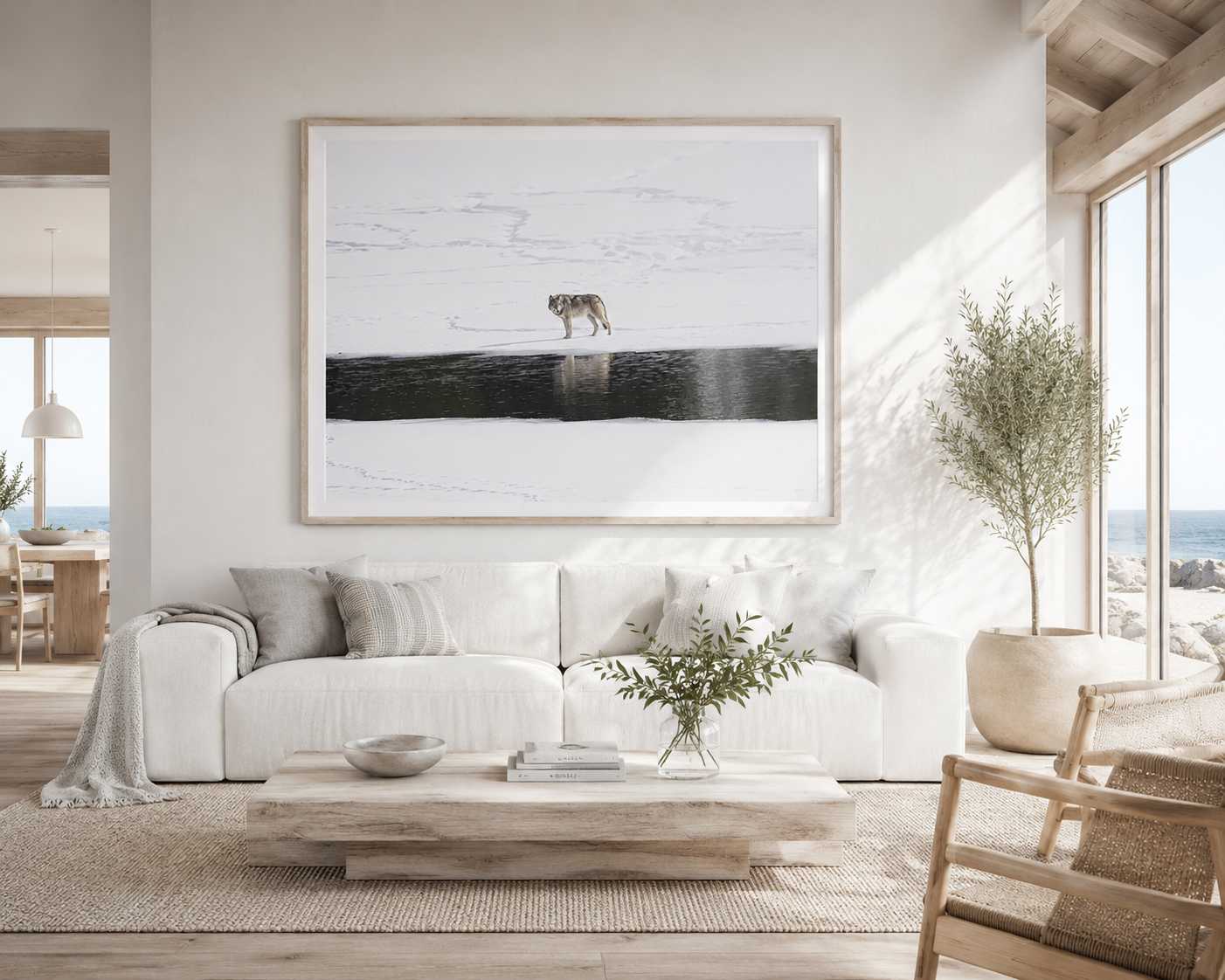 Framed print of a lone wolf standing above dark open water in a snowfield, hung above a linen sectional in a bright, coastal living room