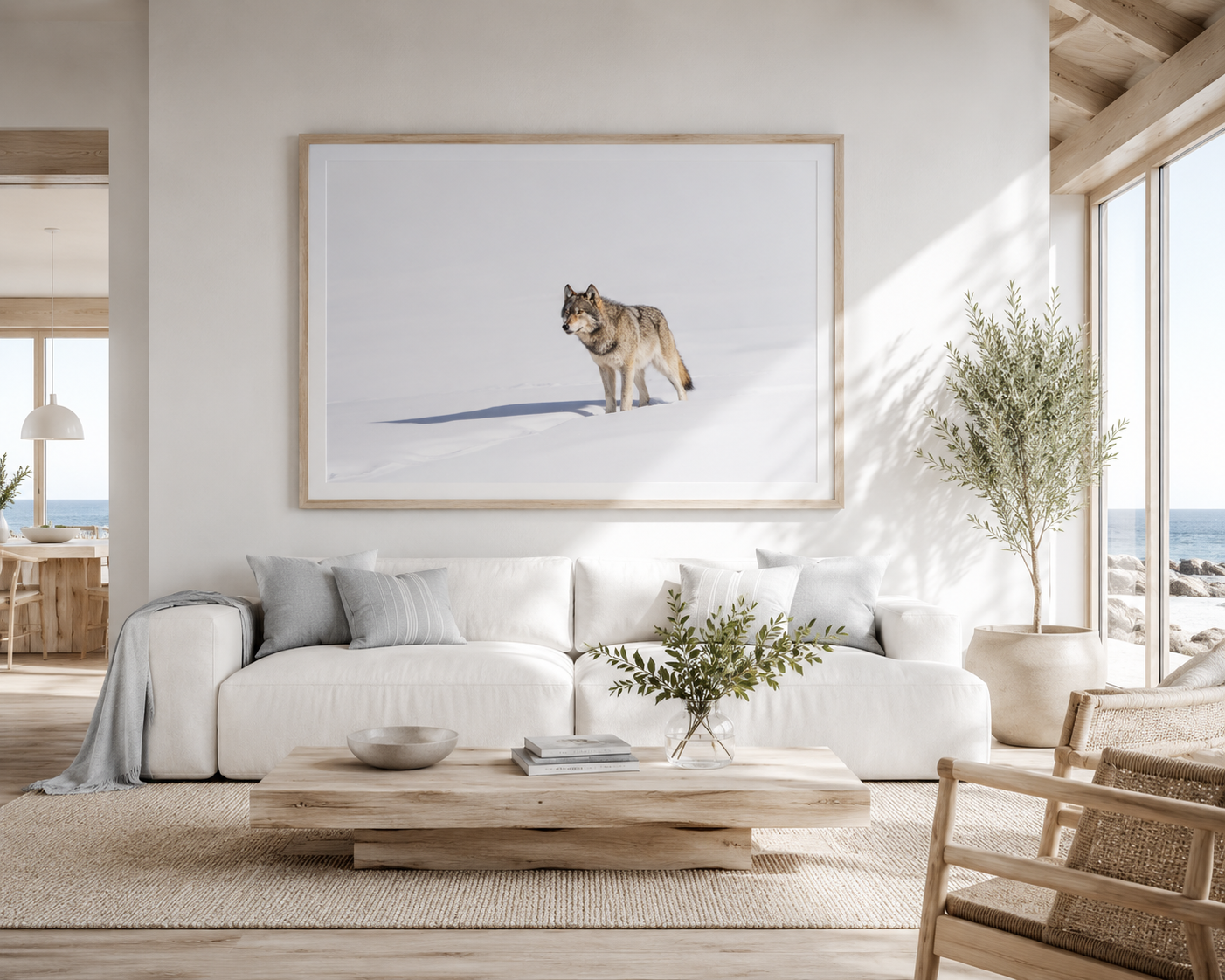 Framed print of a wolf walking through soft-lit snow with her shadow trailing behind, hung above a white sofa in a bright coastal room